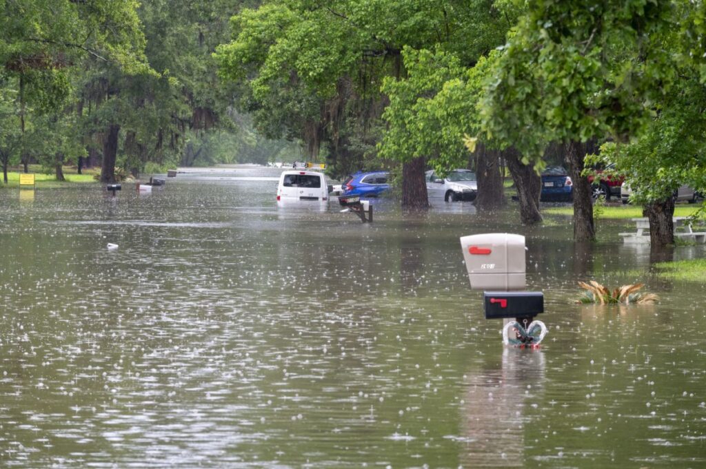 Floodwaters cover a street with cars and mailboxes partially submerged under falling rain.