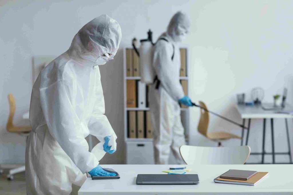 Two people in protective suits sanitize an office, one wiping a desk, the other spraying.