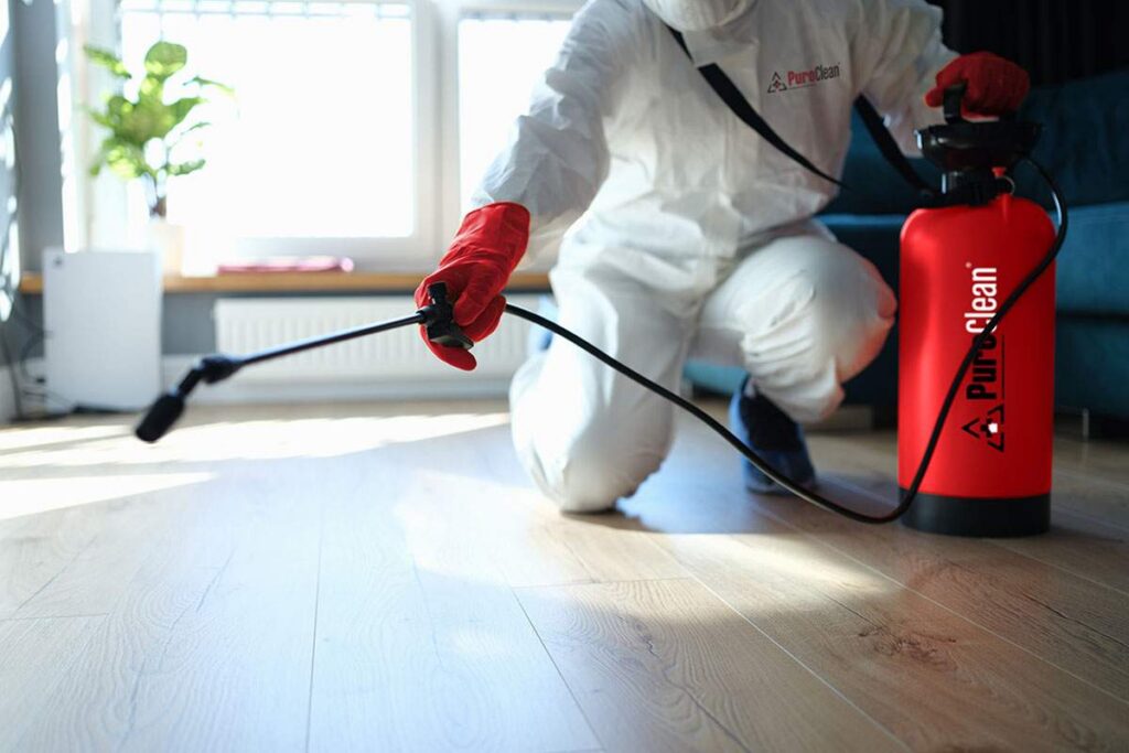 A person in a white protective suit and red gloves sprays a wooden floor with a red PuroClean
