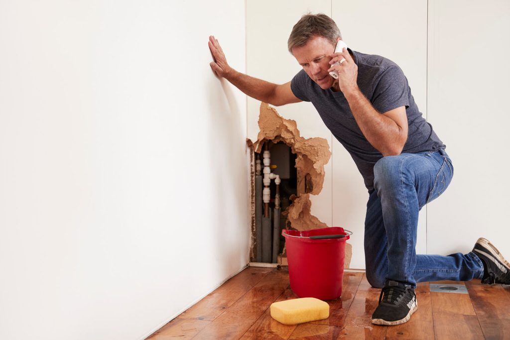 A concerned man kneels, calling on his phone next to a wall with a visible burst pipe