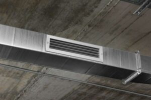 A galvanized metal air duct with a white ventilation grille is mounted on a concrete ceiling