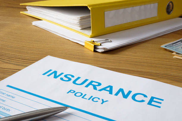 An insurance policy form, pen, yellow binder, and cash are arranged on a wooden desk.