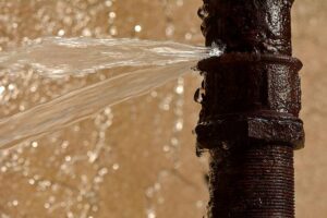 A rusty, dark brown pipe vigorously sprays water from a significant leak. TITLE: