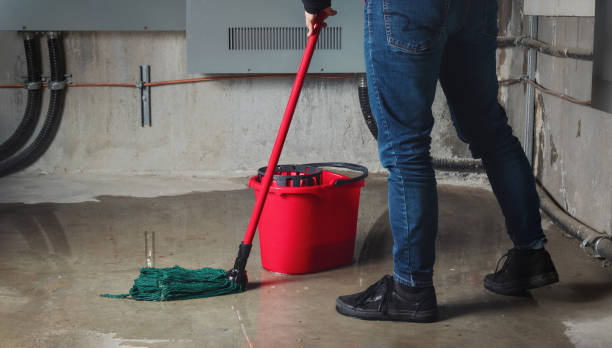 A person in jeans mops a wet concrete floor with a green mop and red bucket
