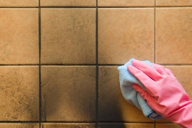 A pink-gloved hand cleans dirty brown tiles with a blue cloth, revealing a clean section.