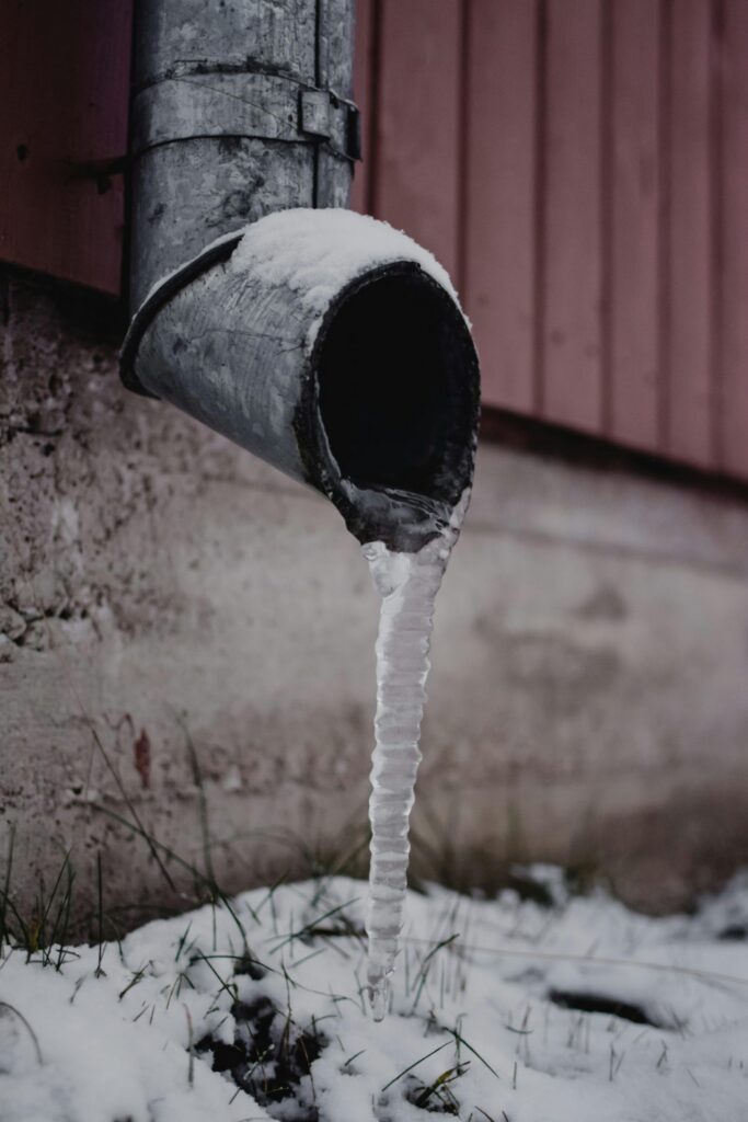 Frozen metal downspout with icicle forming at the base, showing early signs of winter pipe freezing and water damage risk.