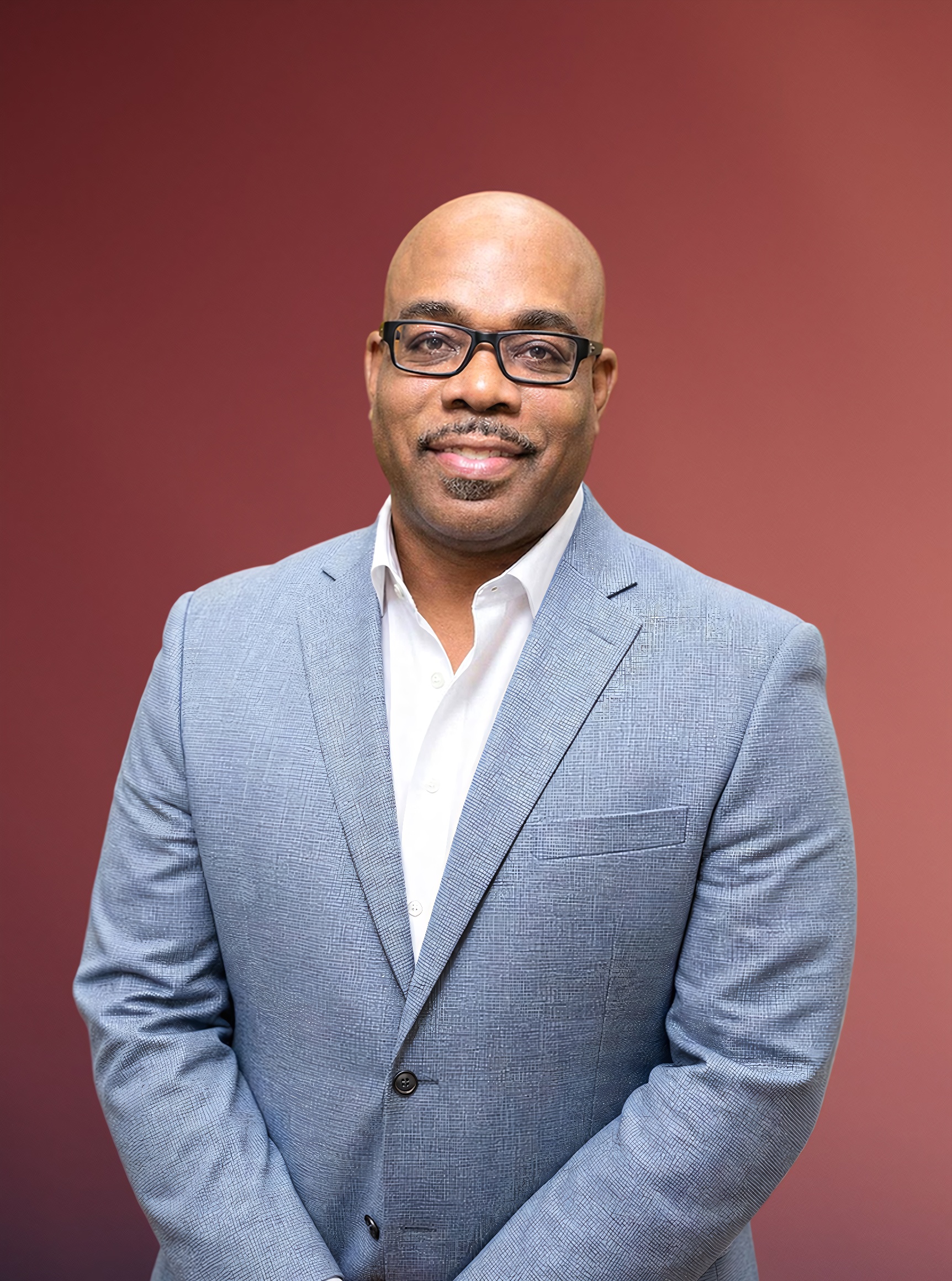 A smiling bald Black man in a light blue blazer and glasses poses against a reddish