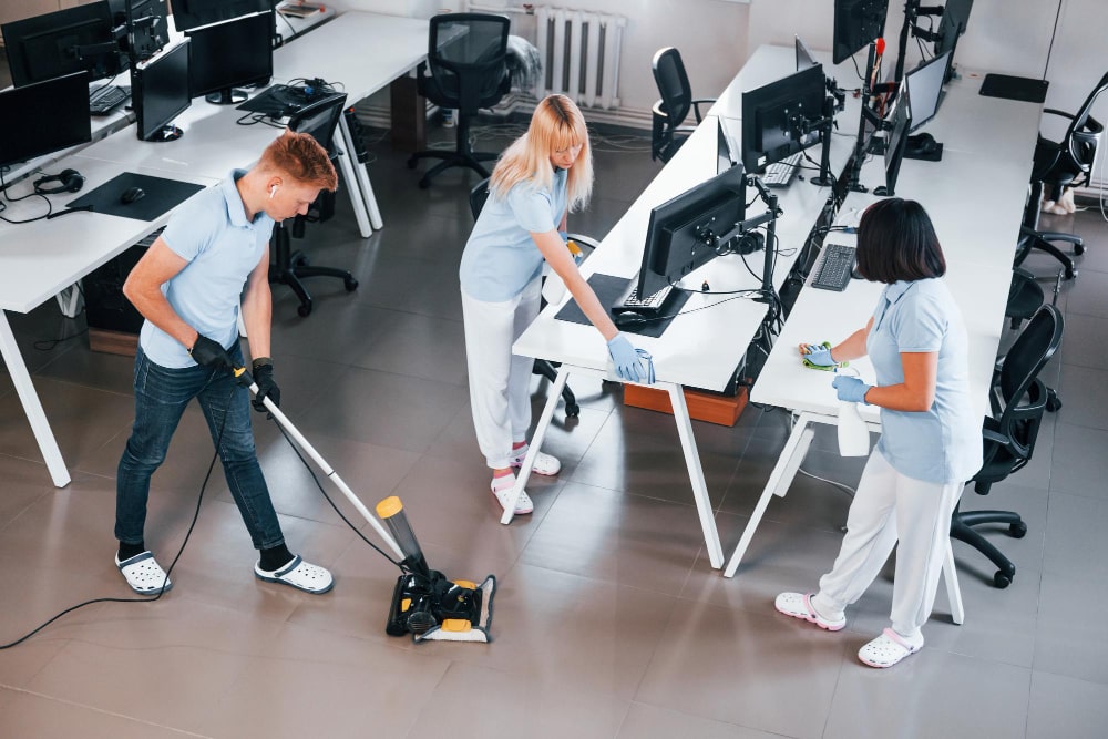 Three professional cleaners tidying an office space with vacuum and cleaning supplies.