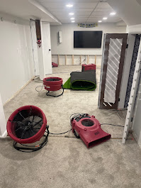Professional water damage restoration in progress in a large basement or commercial space featuring exposed wall framing, concrete floors, industrial air movers, and a high-capacity dehumidifier.