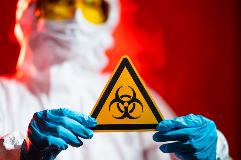 A person in protective gear holds a biohazard warning sign against a red background.