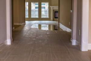 A tiled floor in a house is covered with standing water, reflecting windows and a fireplace.