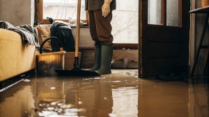 Person wearing rubber boots and gloves cleaning a flooded room after water damage using a broom and bucket.