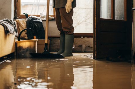 Person wearing rubber boots and gloves cleaning a flooded room after water damage using a broom and bucket.