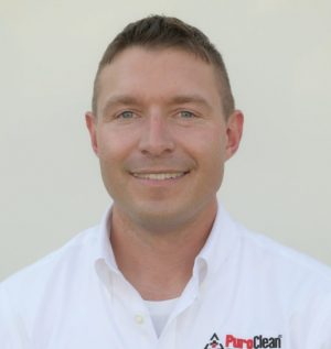 A man wearing a white PuroClean button-up shirt smiles directly at the camera against a plain background.