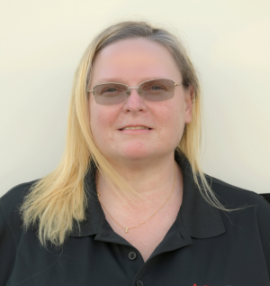 A woman with straight, shoulder-length blonde hair is wearing glasses and a black collared shirt with the "PuroClean" logo on the right side. She is standing in front of a neutral, light-colored background, smiling slightly at the camera.