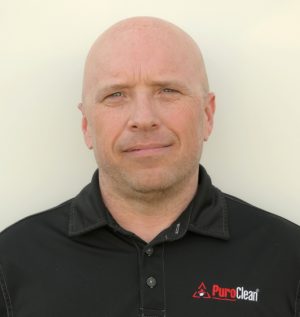 A man wearing a black PuroClean polo shirt stands in front of a plain background, looking directly at the camera.