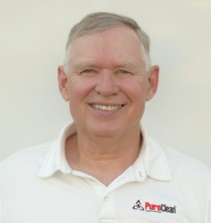 An older man wearing a white PuroClean polo shirt smiles directly at the camera against a plain background.