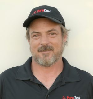 A man with a beard wearing a black collared PuroClean shirt and a black PuroClean cap, smiling at the camera.