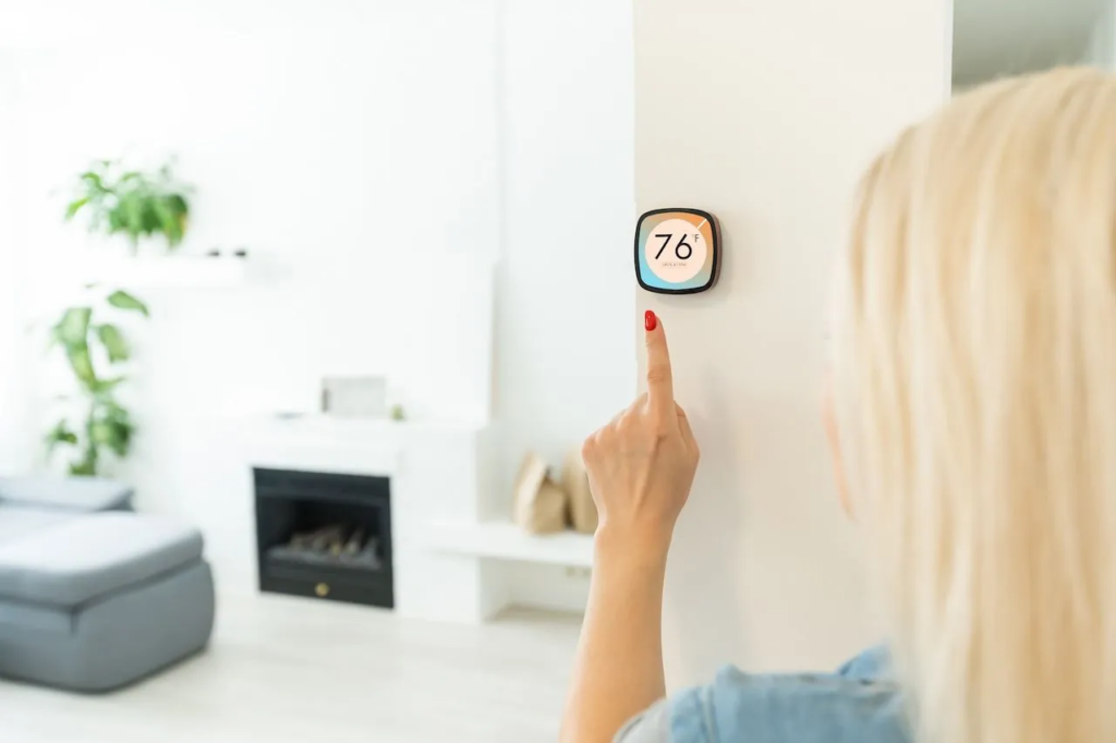 A blonde woman adjusts a smart thermostat displaying 76°F in a modern living