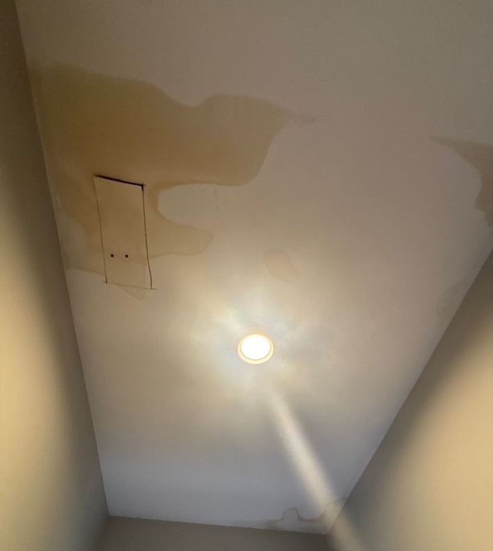 An upward-angled shot of a hallway ceiling showing a severe water stain spreading across the surface. The large, yellowish-brown water stain has distinct, irregular rings and covers a significant portion of the upper left side of the ceiling, surrounding a small rectangular access panel. A glowing, recessed circular light fixture in the center casts a bright glare downward, while smaller, fainter patches of water stain discoloration are visible along the upper right edges and corners of the ceiling.