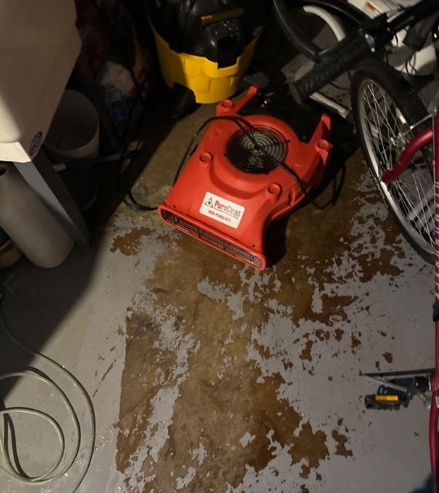 PuroClean red air mover drying standing water on a concrete floor caused by an appliance leak in a residential utility space.