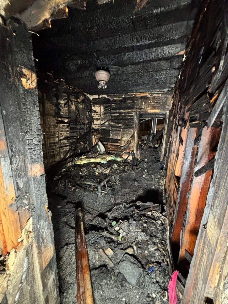 Completely charred room showing severe damage from an electrical fire with melted ceiling fan fixture and exposed wooden framing.