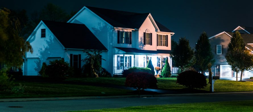 A well-lit suburban house at night, surrounded by trees and neatly trimmed bushes.