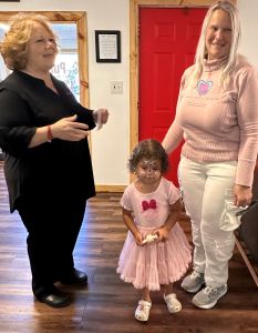 Two women and a young girl in a pink dress stand together, smiling in a bright room with a red door.