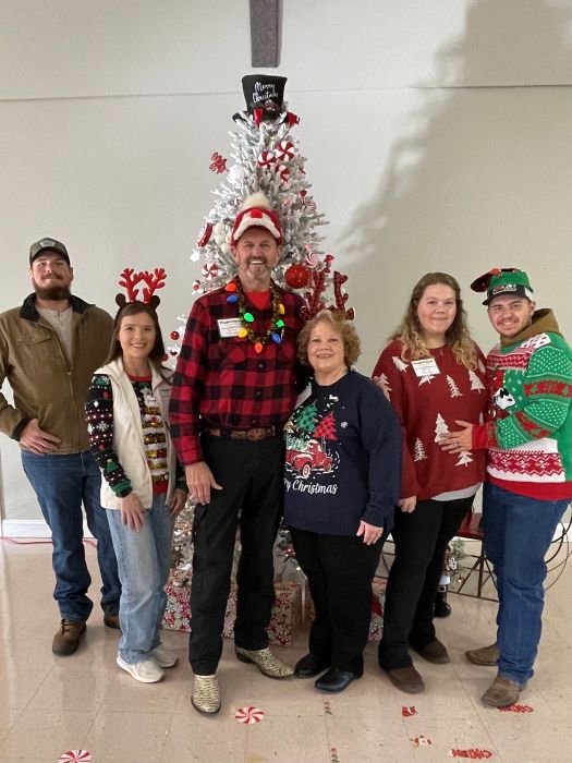 A group of six people in festive attire stands in front of a decorated Christmas tree, smiling and celebrating.