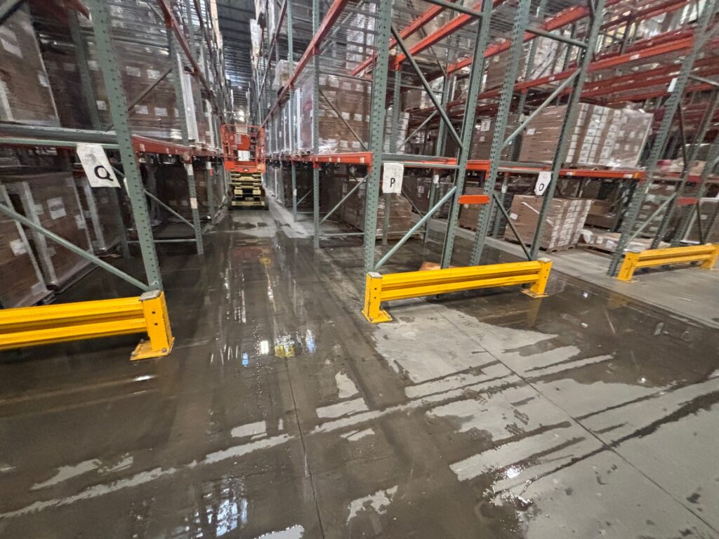 An indoor wide-angle shot of a warehouse aisle floor flooded with water following a pipe burst, reflecting the industrial racking and an orange scissor lift in the background.