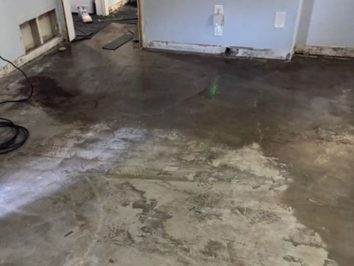 A concrete subfloor showing signs of water damage, discoloration, and damp patches from minor leaks, with removed baseboards and exposed wall framing in the background during a home renovation.