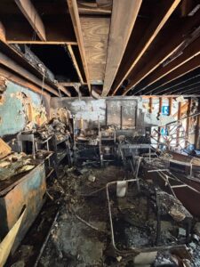Severely charred room showing heavy fire damage to wood ceiling joists and exposed electrical wiring hanging from above.