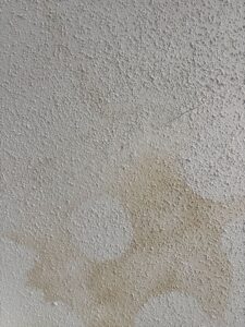 A close-up view of a textured popcorn ceiling showing clear signs of moisture damage. A large, irregular, light-yellowish water stain spreads across the bumpy, white stippled surface. A thin, diagonal hairline crack cuts through the texture, running directly above the stained area.
