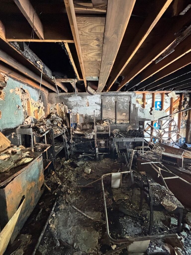 Severely charred room showing heavy fire damage to wood ceiling joists and exposed electrical wiring hanging from above.