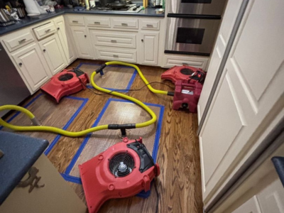 Water damage restoration in progress in a residential kitchen, with red air movers and yellow hoses set up to dry hardwood floors after a leak.