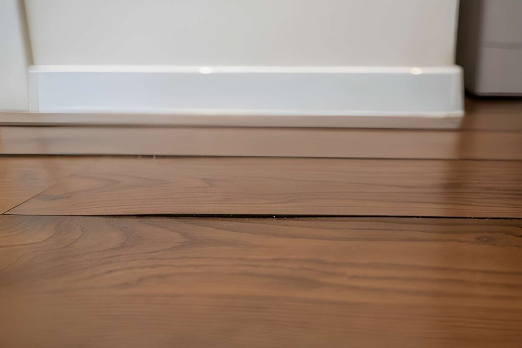 Damaged hardwood floor showing a raised plank and wide gap against a white wall.