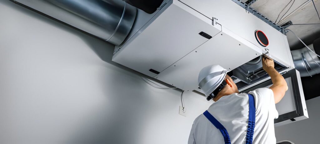 A technician in a hard hat services a large ceiling-mounted ventilation unit. TITLE: