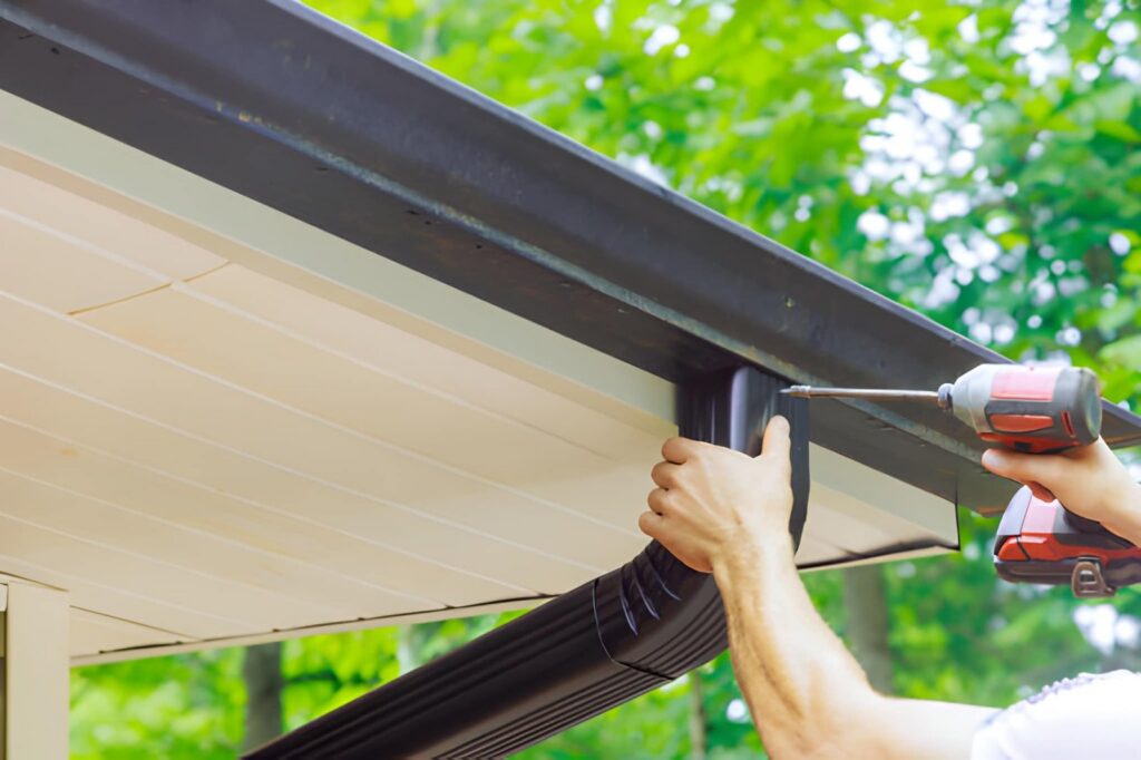 A person installs a black gutter downspout elbow onto a house fascia with a power drill.