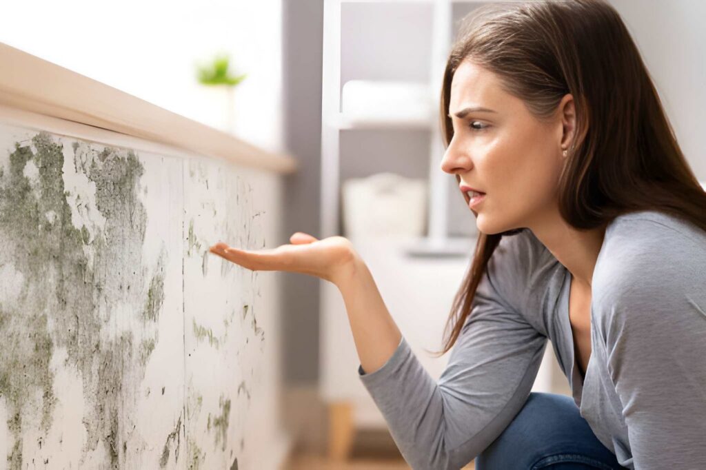 A worried woman observes severe green mold covering a white wall.