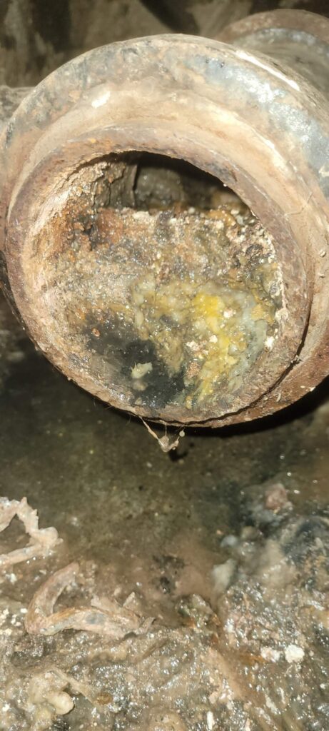 A rusty, corroded pipe is heavily clogged with thick, greasy yellow and grey sludge