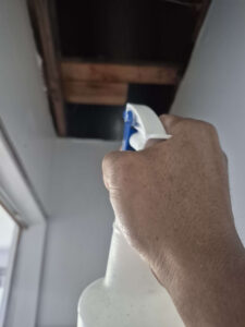 A hand holds a white spray bottle aiming towards an open ceiling access panel.
