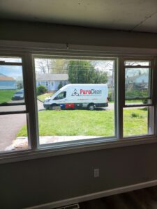 A PuroClean property damage restoration van is parked on a green lawn outside a window in