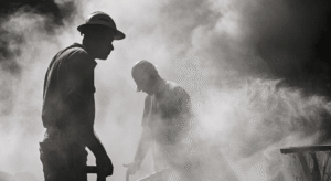 Silhouetted workers in hard hats are busy in a dusty, industrial environment.