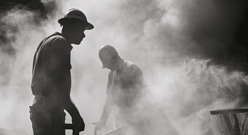 Silhouetted workers in hard hats are busy in a dusty, industrial environment.