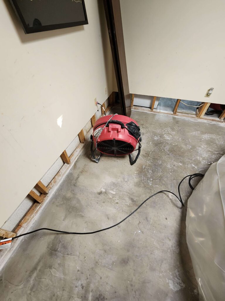 Red industrial fan drying a room with exposed wall studs and a concrete floor. TITLE: