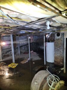 A dimly lit, flooded basement with exposed utilities, insulation, and debris on the wet concrete floor.