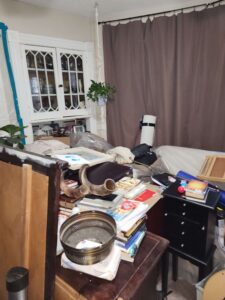 A messy room featuring a white hutch, brown curtains, and a table cluttered with books and