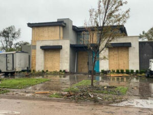 A modern house with many windows boarded up stands on a muddy, debris-strewn lawn after