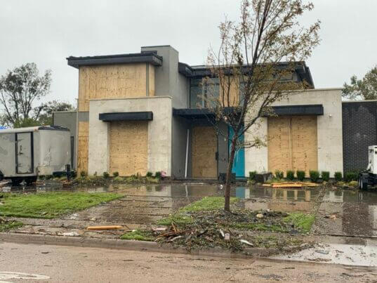 A modern house with many windows boarded up stands on a muddy, debris-strewn lawn after