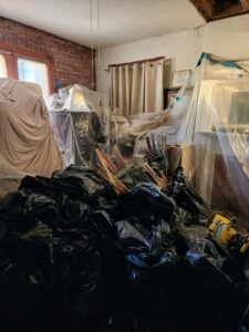 A room undergoing renovation is filled with black trash bags and plastic-covered furniture.